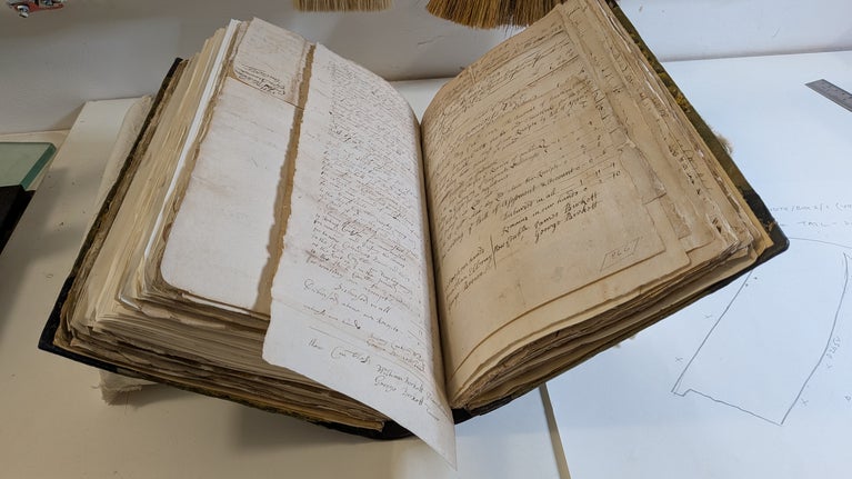 An old handbound book with documents in lies open on a stand on a table.